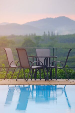 Armchairs with round table at balcony to outdoor background of green natural forest and mountain, with reflection in the swimming poolの写真素材