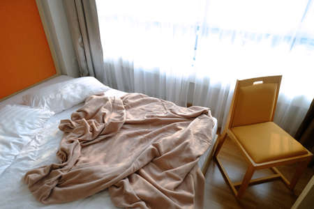 Unmade bed in a bedroom, king size, closed to the window behind the curtain and a comfortable chairの写真素材