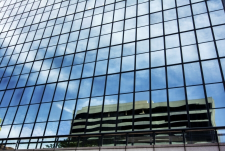 A skyscraper with glass windows refelcting an adjacent building and a beautiful blue sky with white clouds  の写真素材