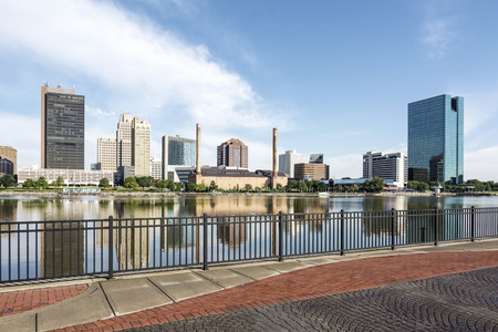 A  panoramic view of downtown Toledo Ohio's skyline from across the Maumee river at a popular restaurant area with a paver brick boardwalk and a decorative iron railing..  A beautiful  blue sky with white clouds for a backdrop.の写真素材