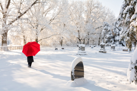 A beautiful winter snow scene with a woman walking in a cemetery with a red umbrella as the snow clings to the trees.の写真素材