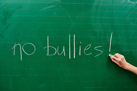 Right hand of a young girl writing the words NO BULLIES on the green school board with a white chalk backgroundの写真素材