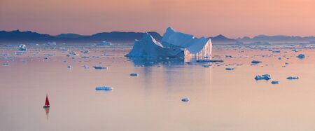 A small boat among icebergs. Sailboat cruising among floating icebergs in Disko Bay glacier during...