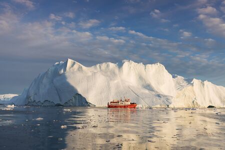 A small boat among icebergs. Sailboat cruising among floating icebergs in Disko Bay glacier during midnight sun Ilulissat, Greenland. Studying of global warming phenomenon Ices and icebergsの写真素材