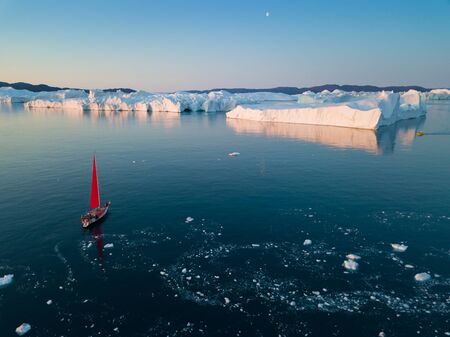 Little Red Sailboat Cruising Among Floating Icebergs In Disko Bay Glacier During Midnight Sun Season...
