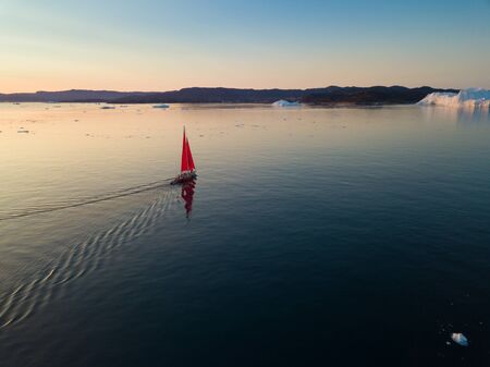 Little Red Sailboat Cruising Among Floating Icebergs In Disko Bay Glacier During Midnight Sun Season...