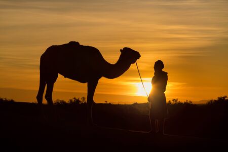Bedouin and camel on the desert through the desert Beautiful sunset with caravan on Sahara, Morocco Desert with camel and nomads Silhouette nomad man with dramatic sky Picturesque background nature conceptの写真素材
