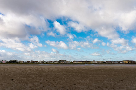 Wide, layered view of Sandymount Beach, Dublin. A vast expansion of low tide sand leads to a long, uniform line of classic seaside terrace houses under a dynamic blue and cloudy sky.の写真素材