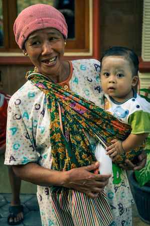 Yogyakarta in Java, Indonesia - May 2015: Smiling woman with pink cap wears colourful scarf with small black-haired child with white powder on face in it in Yogyakarta, city in Java, Indonesia. Documentary editorial.のeditorial素材