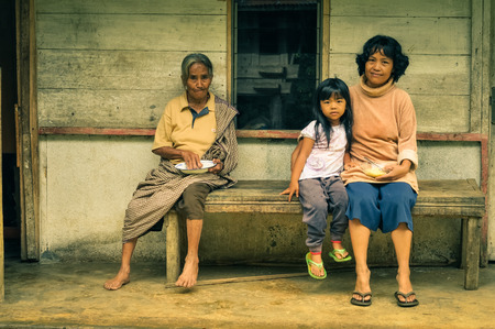 Toraja, Indonesia - June 2015: Grandmother sits on bench in front of her house with her daughter and granddaughter and they hold plates and look to photocamera in Toraja, Sulawesi region, Indonesia. Documentary editorial.のeditorial素材