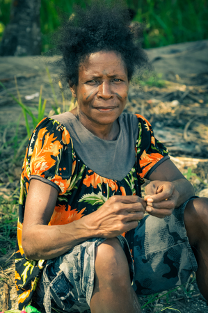 Aitape, Papua New Guinea - July 2015: Native woman with black hair dressed in colourful shirt sits on ground and looks to photocamera in Aitape, Papua New Guinea. Documentary editorial.のeditorial素材