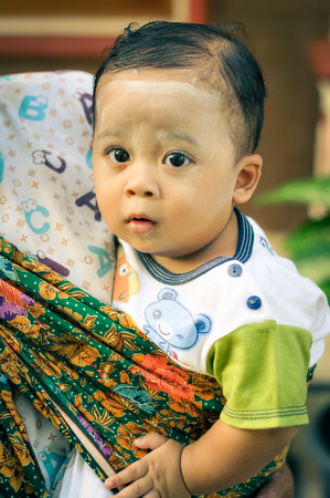 Yogyakarta in Java, Indonesia - May 2015: Small black-haired child with white powder on face looks curiously to photocamera with big black eyes from arms of his mother in Yogyakarta, city in Java, Indonesia. Documentary editorial.のeditorial素材