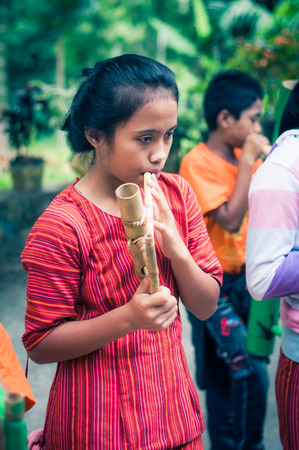 Suaya, Indonesia - May 2015: Young black-haired girl dressed in red shirt with stripes plays bamboo flute in Suaya, in the south-east of Tana Toraja, Sulawesi, Indonesia. Documentary editorial.のeditorial素材