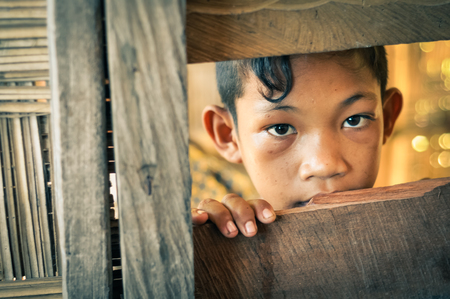 Malange, Indonesia - June 2015: Young native boy with nice dark brown eyes looks to photocamera from hole in wooden wall in Malange, Togean islands, Sulawesi, Indonesia. Documentary editorial.のeditorial素材