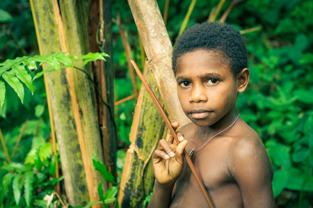 Will-will, Papua New Guinea - July 2015: Young half-naked boy stands in forest with wooden stick in hands and looks to photocamera in background with beautiful greenery of forest in Will-will, Nuku, Papua New Guinea. Documentary editorial.のeditorial素材