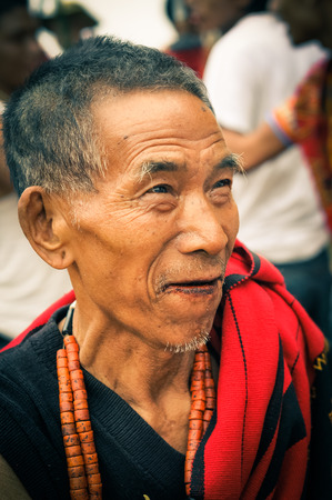 Wanching, Nagaland - April 2012: Older man with orange necklace made of beads and red scarf looks up at Aoleang festival in Wanching, Nagaland. Documentary editorial.のeditorial素材