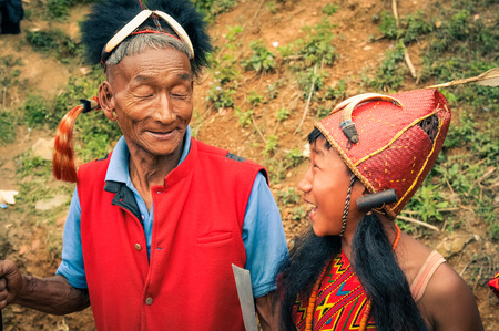 Mon, Nagaland - April 2012: Older native man in red vest and with hat made of fur and feather at top poses with young boy in traditional costume at Aoleang festival in Mon, Nagaland. Documentary editorial.のeditorial素材