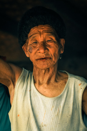 Wanching, Nagaland - April 2012: Old man with wrinkled face wears white t-shirt and looks to photocamera during Aoleang festival in Wanching, Nagaland. Documentary editorial.のeditorial素材