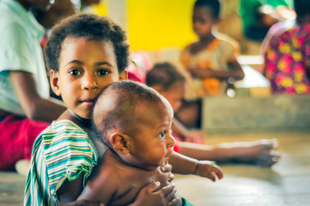 Palembe, Papua New Guinea - July 2015: Small girl looks to photocamera with big brown eyes with her small sibling in her arms in Palembe, Sepik river, Papua New Guinea. Documentary editorial.のeditorial素材