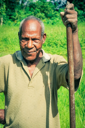 Palembe, Papua New Guinea - July 2015: Older native man in yellow shirt holds wooden stick and poses on green field in Palembe, Sepik river, Papua New Guinea. Documentary editorial.のeditorial素材