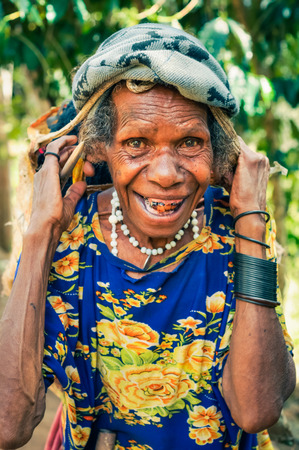 Kubor Range, Papua New Guinea - August 2015: Old native woman in blue and yellow shirt with cap on her head carries load and smiles to photocamera in Kubor Range, Papua New Guinea. Documentary editorial.のeditorial素材