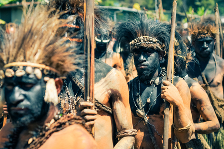 Wabag, Papua New Guinea - August 2015: Native half-naked men with black colour on faces and headbands with feathers hold wooden sticks and walk in row during traditional Enga cultural show in Wabag, capital of Enga Province, Papua New Guinea. Documentary のeditorial素材