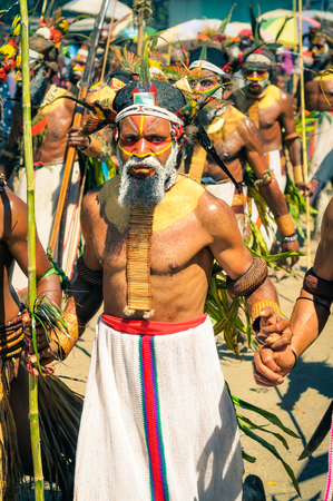 Wabag, Papua New Guinea - August 2015: Native man dressed in traditional white skirt with large necklace, headband and cap with face covered in colours walks with bamboo spear during Enga cultural show in Wabag, capital of Enga Province, Papua New Guinea.のeditorial素材