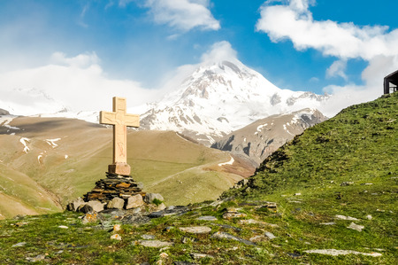 Photo of large cross standing on stones surrounded by greenery of high mountains in Kazbegi, Georgia.の写真素材