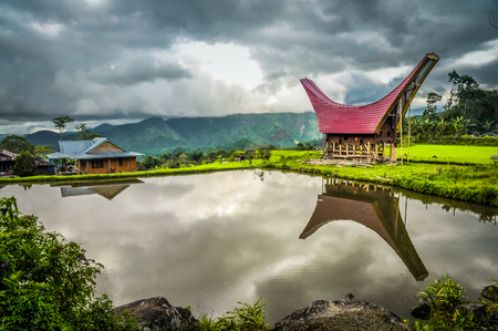 Photo of tongkonan and house near lake with view on mountains in distance in Toraja region in...