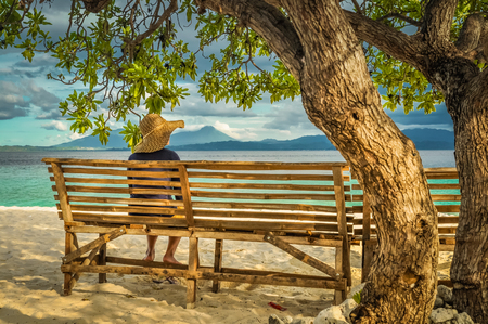 Pulau Lihaga, North Sulawesi, Indonesia - June 2015: Photo of native woman with straw hat sitting on bench under tree on sand beach and watching sea in Pulau Lihaga in North Sulawesi, Indonesia. Documentary editorial.のeditorial素材