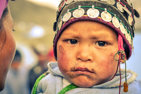 Dolpo, Nepal - circa May 2012: Small child with cap with coins on head has dirt on face and frowns to photocamera with black eyes in Dolpo, Nepal. Documentary editorial.のeditorial素材