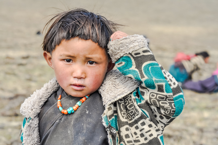 Dolpo, Nepal - circa June 2012: Small native girl with short black hair dressed in warm blue and black jacket with colourful necklace touches her head with hand in Dolpo, Nepal. Documentary editorial.のeditorial素材