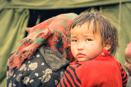 Dolpo, Nepal - circa June 2012: Small brown-haired girl in red sweater looks over shoulder to photocamera with brown eyes in Dolpo, Nepal. Documentary editorial.のeditorial素材