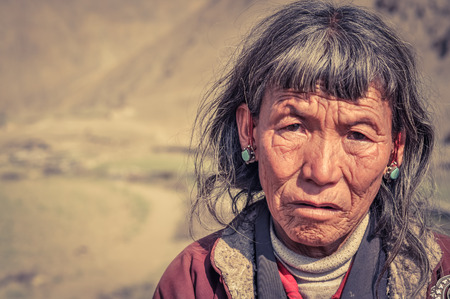 Dolpo, Nepal - circa June 2012: Old grey-haired woman with wrinkled face wears blue earrings and looks sadly to photocamera in Dolpo, Nepal. Documentary editorial.のeditorial素材