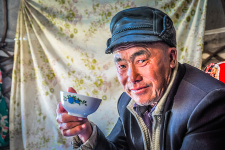 Son Kol, Kyrgyzstan - circa September 2011: Older man with leather cap poses during drinking of tea in nomad tent in Son Kol, Kyrgyzstan. Documentary editorial.のeditorial素材