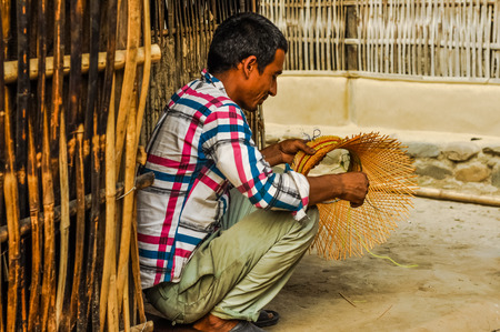 Damak, Nepal - circa May 2012: Native man in checked blue and red shirt kneels and weaves hat outside at Nepali refugee camp in Damak, Nepal. Documentary editorial.のeditorial素材