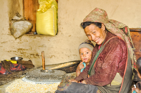 Dolpo, Nepal - circa May 2012: Smiling woman with headcloth with child and sits on ground near stone hand grain mill in Dolpo, Nepal. Documentary editorial.のeditorial素材