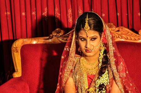 Dhaka, Bangladesh - circa July 2012: Young black-haired bride in beautiful red and gold glittering dress sits on red couch and looks right in Dhaka, Bangladesh. Documentary editorial.のeditorial素材