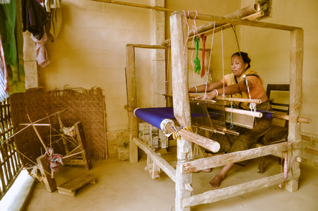 Srimongal, Bangladesh - circa July 2012: Barefoot black-haired woman with yellow scarf sits on wooden chair and weaves with blue cloth in Srimongal, Bangladesh. Documentary editorial.のeditorial素材