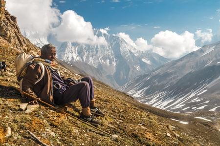Dolpo, Nepal - circa June 2012: Native man with bag on his back and wooden stick in his hand sits on ground and rests in beautiful snowy mountains in Dolpo, Nepal. Documentary editorial.のeditorial素材