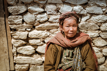 Dolpo, Nepal - circa May 2012: Native woman with red headcloth and scarf wears brown jacket and sits in front of stone house in Dolpo, Nepal. Documentary editorial.のeditorial素材