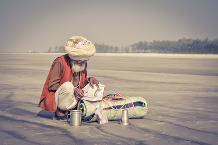 Ganga Sagar, West Bengal - circa January 2012: Native man with white turban squats and reads book in Ganga Sagar, West Bengal. Documentary editorial.のeditorial素材