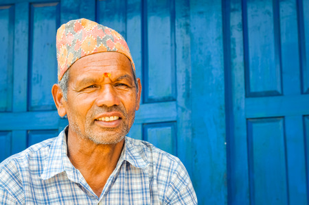 Beni, Nepal - circa May 2012: Old man with colourful cap on head and red dot on forehead wears white shirt with big blue door behind him in streets of Beni, Nepal. Documentary editorial.のeditorial素材