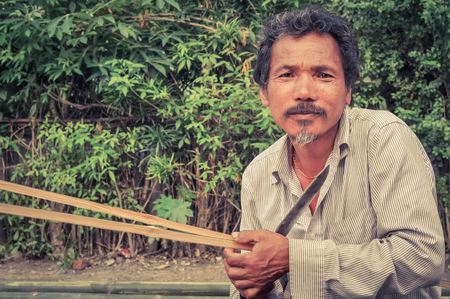 Damak, Nepal - circa May 2012: Old kneeling man dressed in white clothes holds knife at Nepali refugee camp in Damak, Nepal. Documentary editorial.のeditorial素材