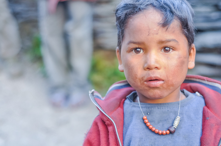 Dolpo, Nepal - circa May 2012: Young boy with brown hair and eyes has dirt on cheeks and wears beads and grey shirt with red sweatshirt in Dolpo, Nepal. Documentary editorial.のeditorial素材