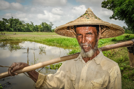 Srimongal, Bangladesh - circa July 2012: Old man with wrinkles on face and with grey beard holds wooden stick on his shoulder in Srimongal, Bangladesh. Documentary editorial.のeditorial素材