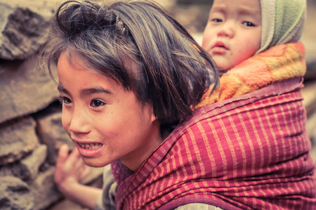 Old Dirang, Arunachal Pradesh - circa February 2012: Young girl with brown eyes and curious sight carries her younger sibling on her back in Old Dirang, Arunachal Pradesh. Documentary editorial.のeditorial素材