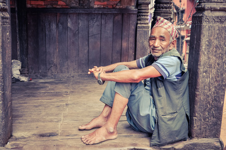 Bhaktapur, Nepal - circa June 2012: Native man in blue clothes and cap on head sits on ground and leans on wooden column in Bhaktapur, Nepal. Documentary editorial.のeditorial素材