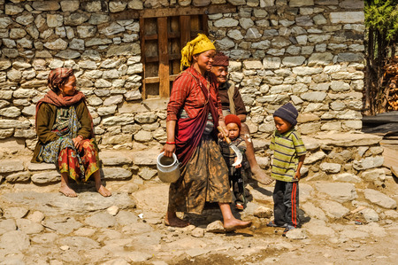 Dolpo, Nepal - circa May 2012: Smiling mother in red clothes walks with her two children and woman with man sit on stone bench in front of stone house in Dolpo, Nepal. Documentary editorial.のeditorial素材