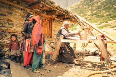 Dolpo, Nepal - circa June 2012: Old grey-haired woman in brown dress weaves on wooden loom outside her stone house and young girls watch her in Dolpo, Nepal. Documentary editorial.のeditorial素材